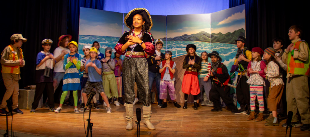 A full cast of students are dressed as pirates in front of a backdrop of an ocean. 