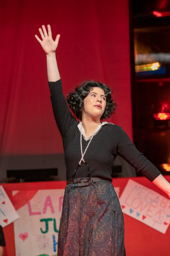 A student in a brown curly wig performs a song on stage with one hand raised in the air. 