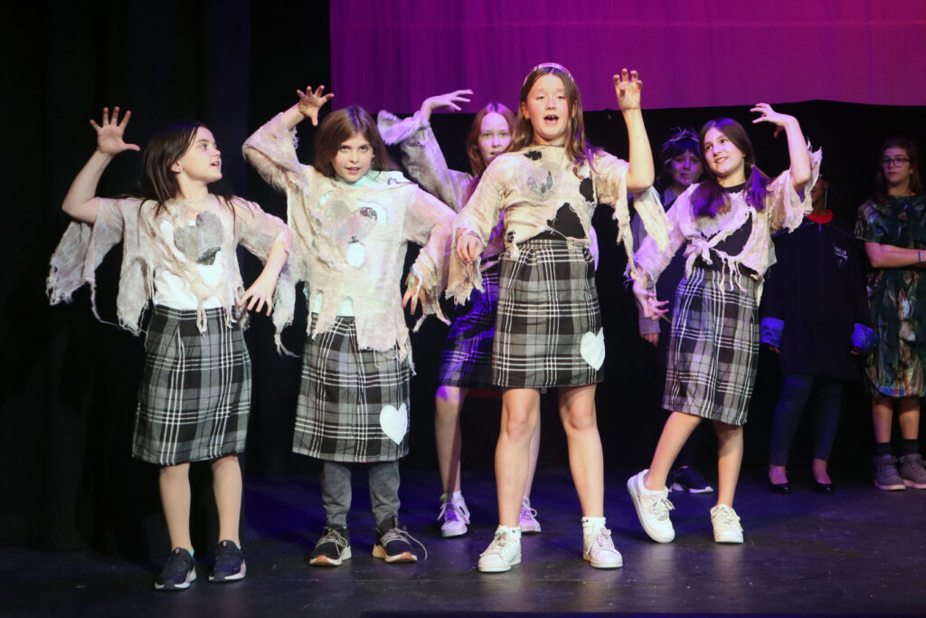 A group of girls dressed as zombies dance on stage with their hands up. 