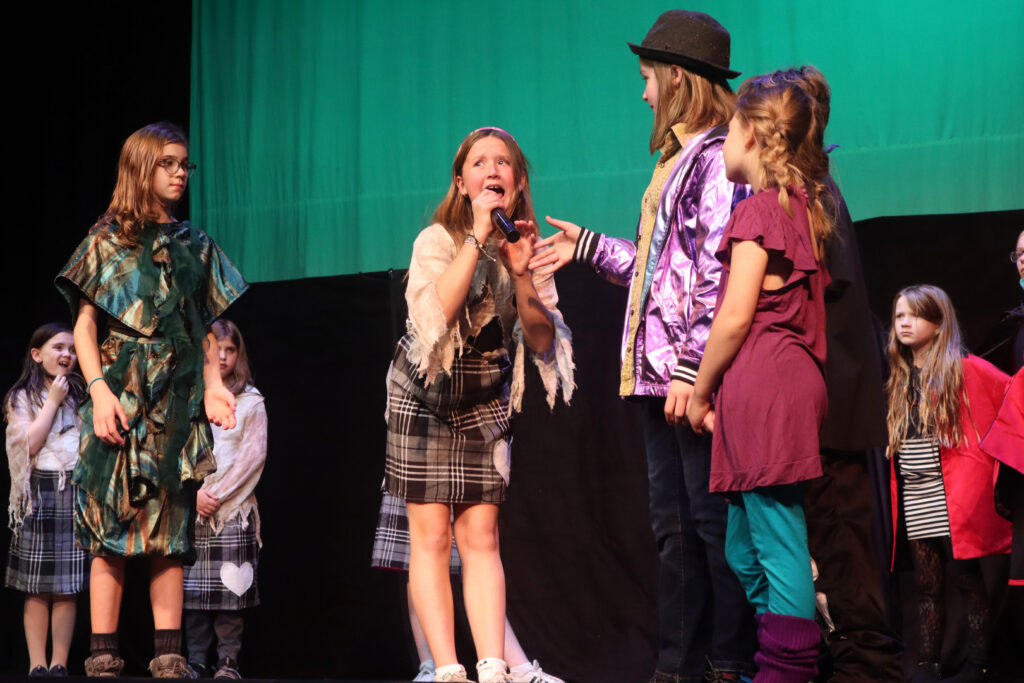 A student grabs a microphone from another actor on stage with a green backdrop behind them. 