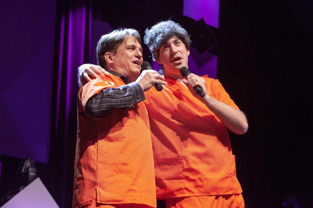 Two men in orange jumpsuits sing and hold onto each others shoulders with mics in their hands. 
