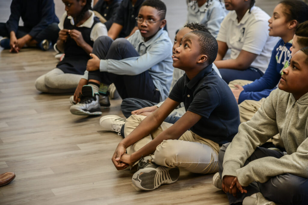 A group of students sit on the floor listening intently to a teacher. 