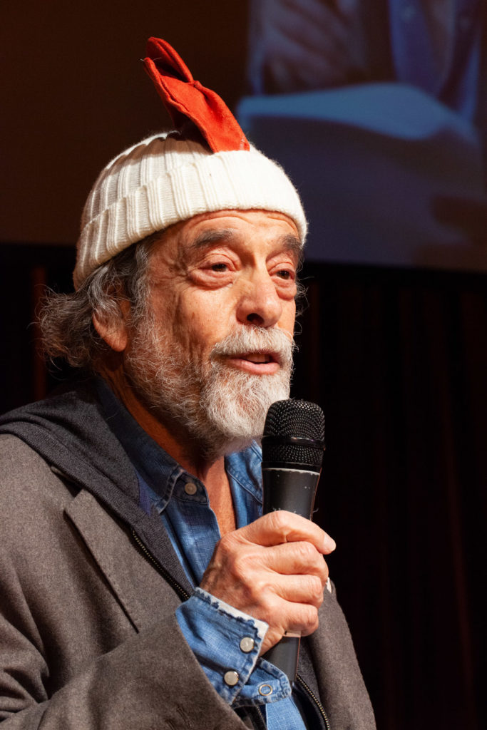 An old man wears a rooster hat and speaks into a microphone. 