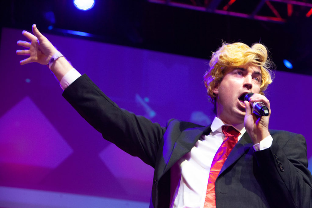 A man in a blonde wig and suit and tie sings into a microphone with his hand outstretched dramatically. 