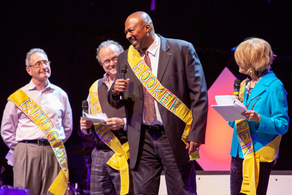 A group of adults stand on the stage with microphones, all wearing yellow sashes.