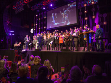 A large group of performers sing on stage with purple and blue lights. 