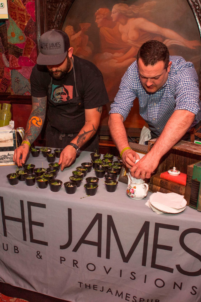 Two chefs prepare food from across a table, leaning down to put food in tiny cups. 