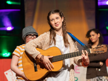 A young performer plays guitar on stage with braids in her hair. 