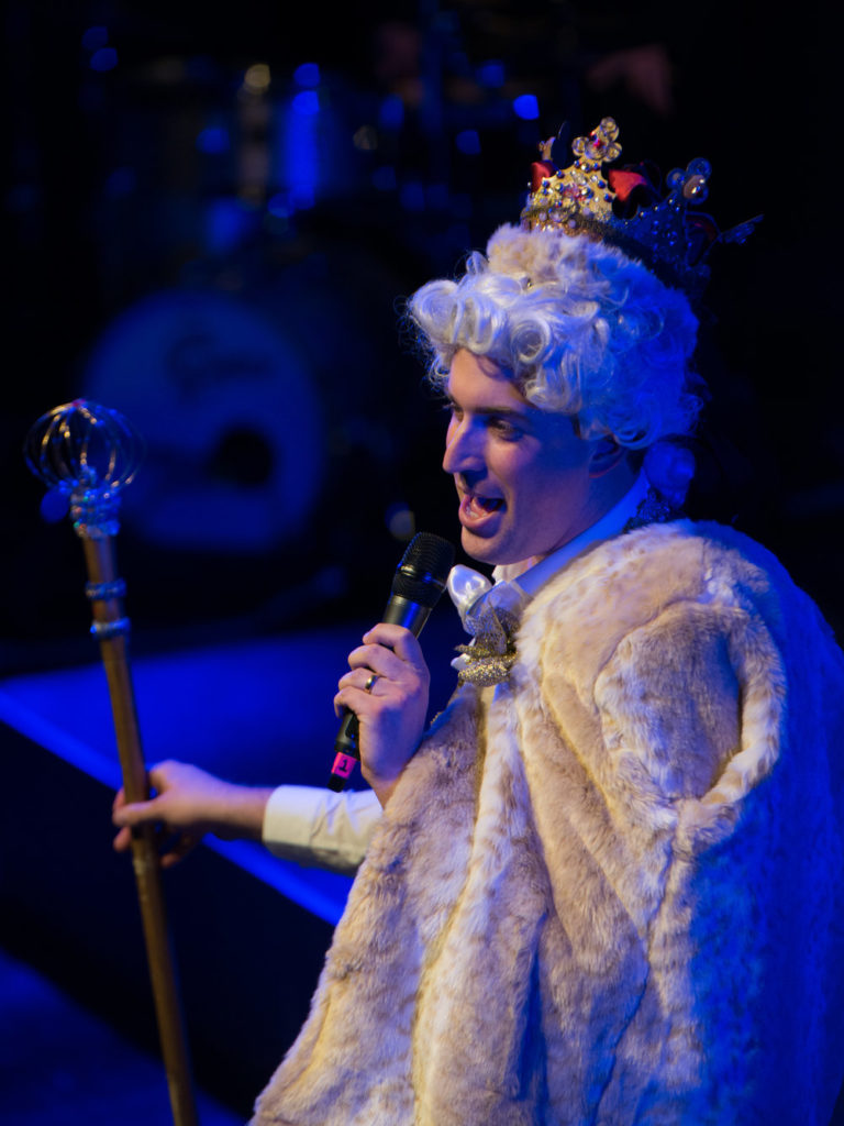 A man dressed as King George, carrying a staff and wearing a crown and cape, sings into a microphone.  