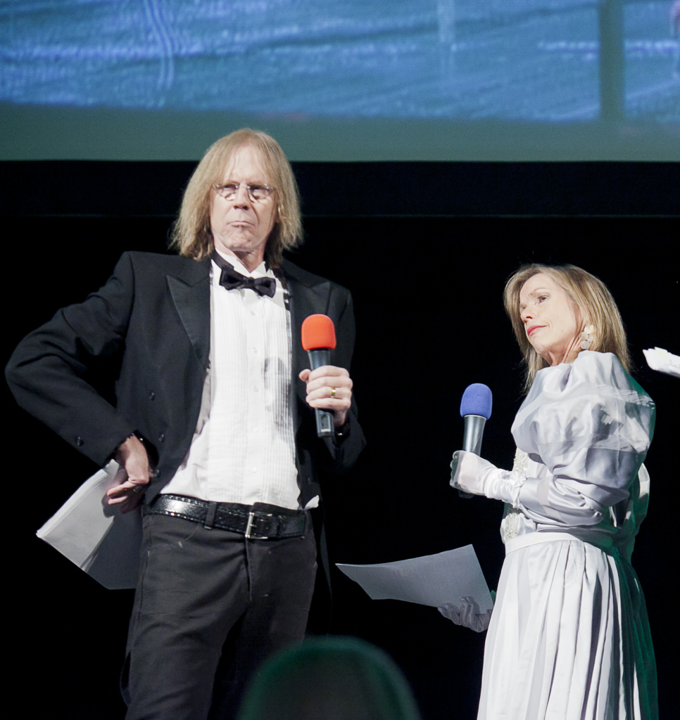 A man and woman stand opposite each other, dressed in a tux and a ballgown, with microphones. 